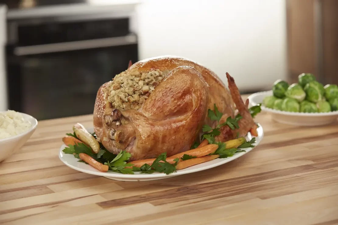 A roasted turkey stuffed with herbs on a platter, surrounded by carrots and greens, on a wooden kitchen counter.