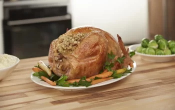 A roasted turkey stuffed with herbs on a platter, surrounded by carrots and greens, on a wooden kitchen counter.